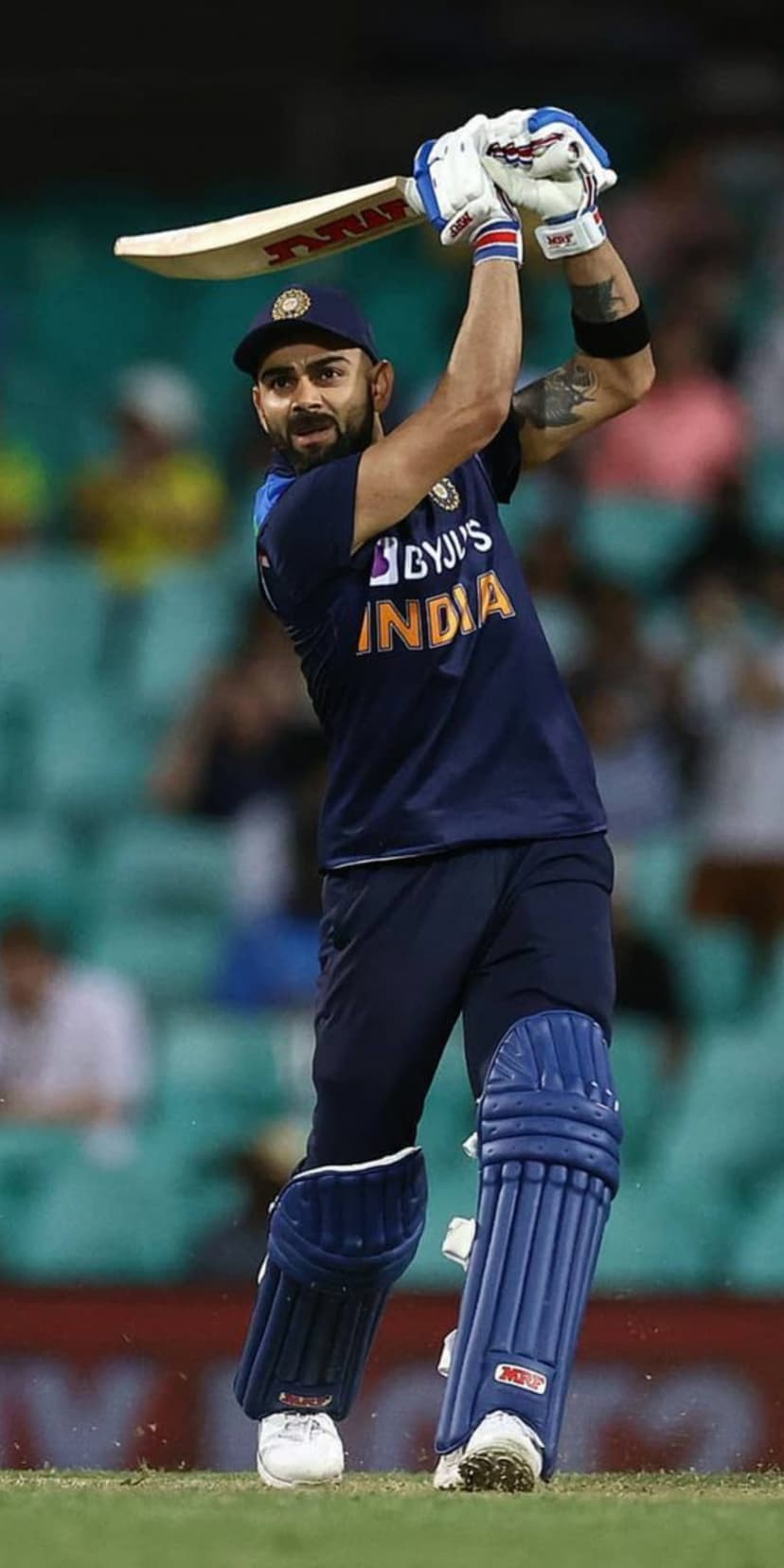 Virat Kohli wearing a blue India jersey swings a cricket bat with a focused expression on a grassy stadium field.