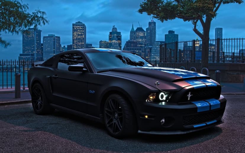 Matte black Ford Mustang with blue racing stripes and halo headlights parked in front of a city skyline at dusk.