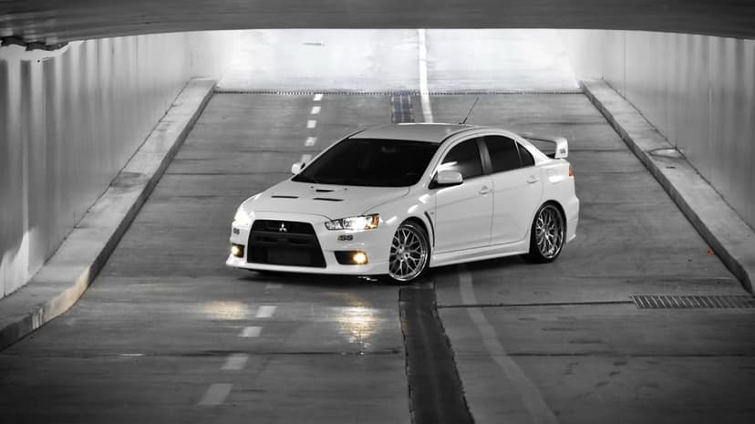 White Mitsubishi Lancer Evolution X with glowing headlights parked in a gray concrete tunnel at an angle.