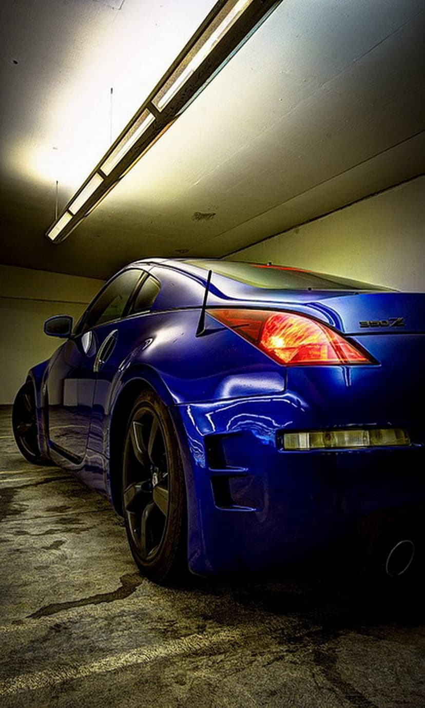 Blue Nissan 350Z with black rims and a rear spoiler parked on a concrete garage floor under overhead light strips.
