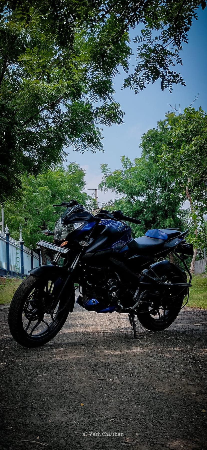 Blue sport bike parked on a gravel road surrounded by thick green trees and a bright clear sky during the day.