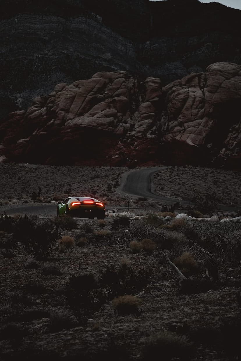 Green Lamborghini with lit taillights drives on a winding desert road between rocky mountains during twilight.