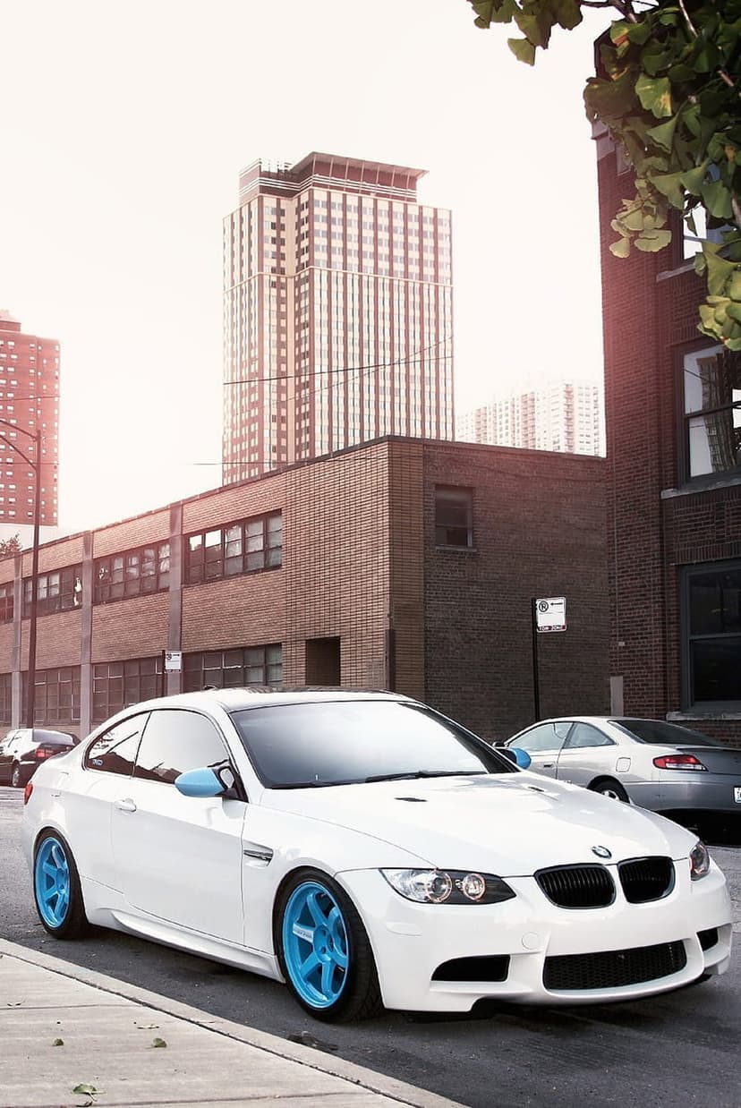 White BMW coupe with bright blue rims parked on a city street between brick buildings and tall skyscrapers.