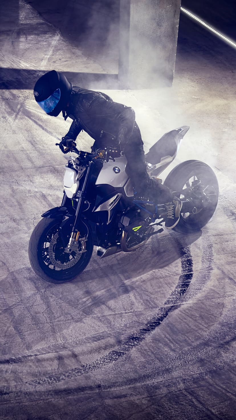Motorcyclist in black leather gear performs a smoky burnout on a white sportbike amidst dramatic studio lighting.