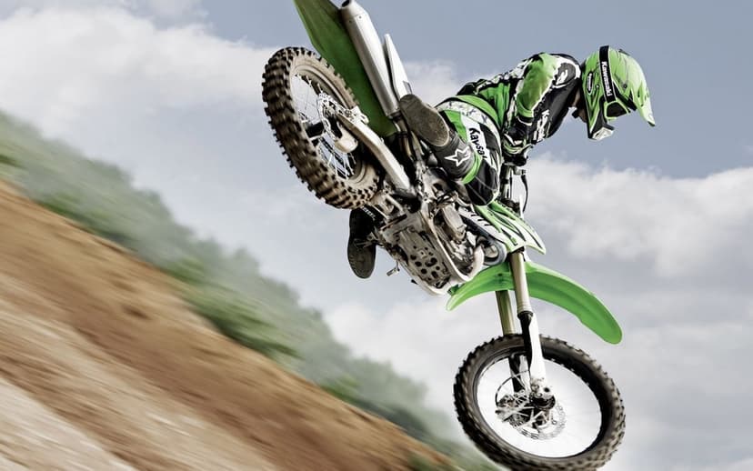 Motocross rider in green gear jumping a Kawasaki dirt bike mid-air over a blurred dirt track and cloudy sky.