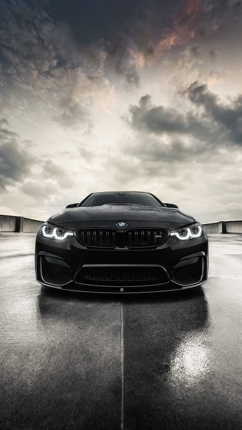 Black BMW M4 with glowing headlights on a wet concrete road under a dark, cloudy sky at dusk.