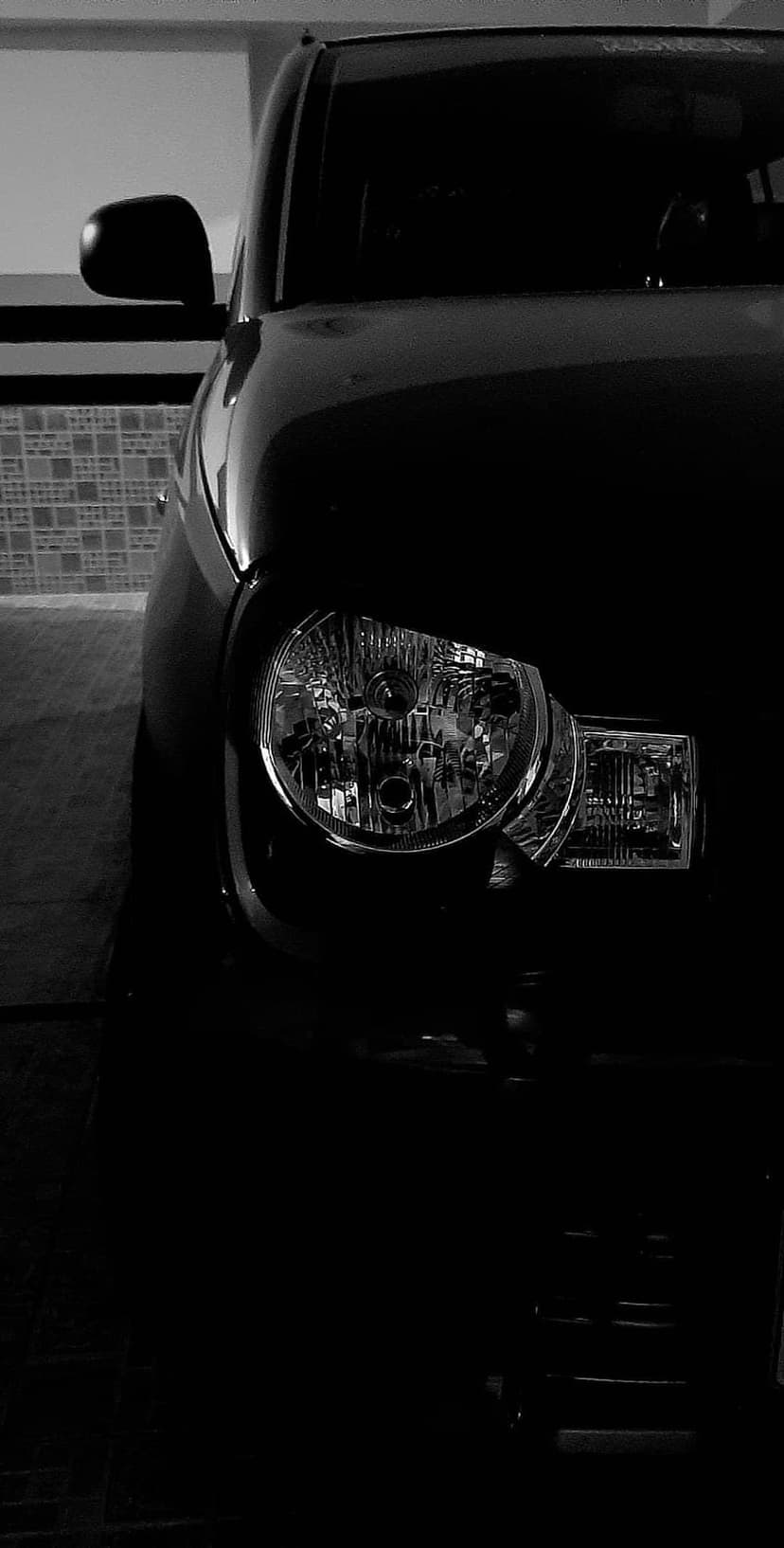 Car headlight assembly and lens reflectors emerge from deep shadows with a side mirror visible in the background.