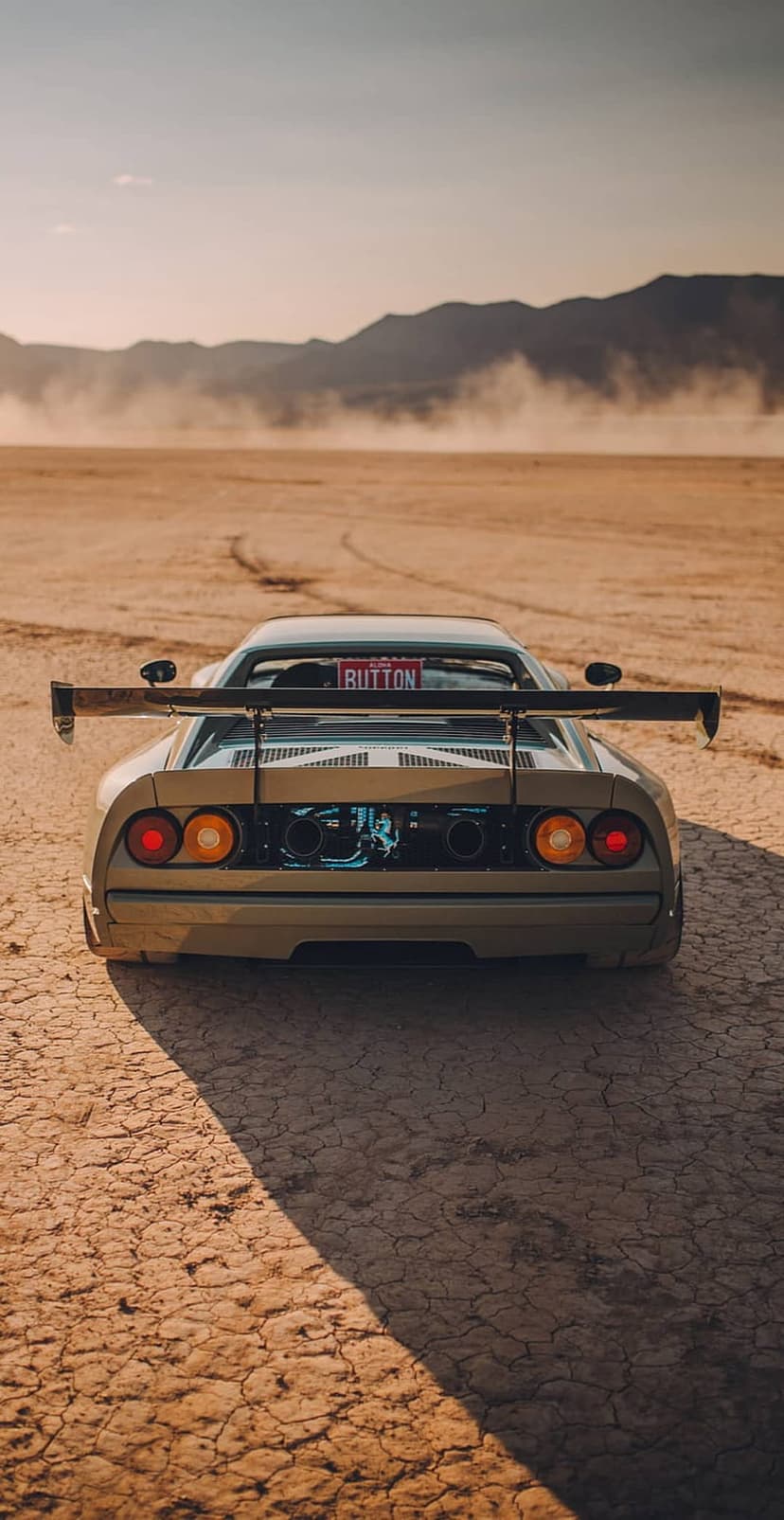 Tan Ferrari F40 rear view speeding through a dusty desert with a custom BUTTON plate and large rear wing.