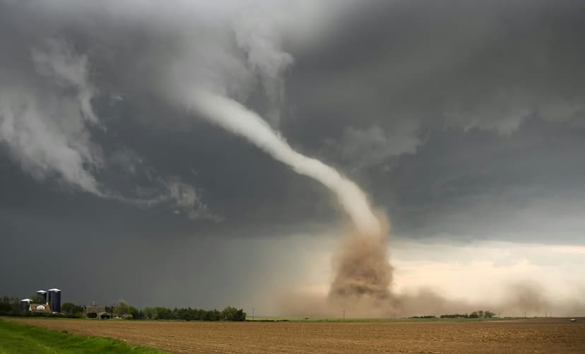 Large funnel cloud touching down on a flat rural field with a distant farmhouse and silo under dark storm clouds.