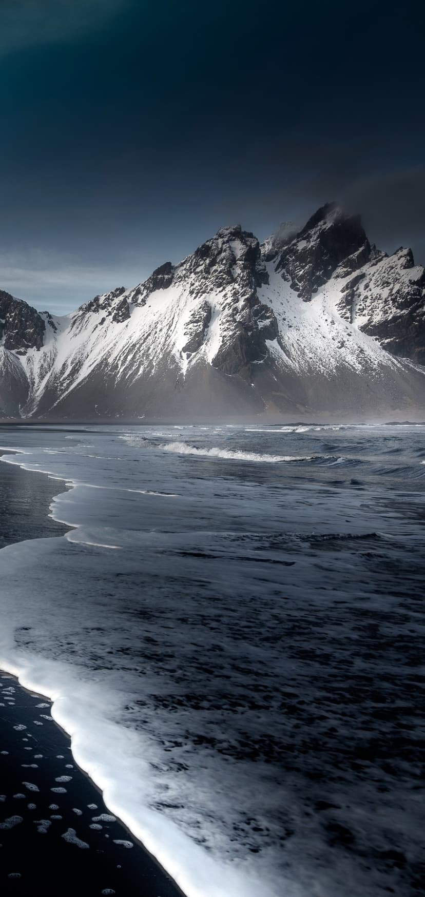 Black sand beach with white foam waves meeting snowy mountains under a dark, moody sky in vertical orientation.