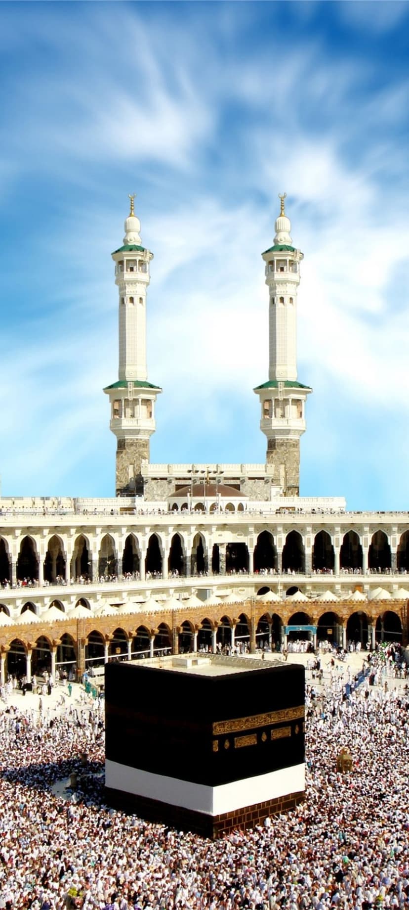 The Kaaba in Mecca surrounded by white-clad pilgrims and mosque minarets under a streaked, cloudy sky.