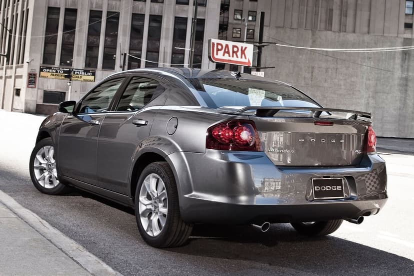 Grey Dodge Avenger sedan rear view parked on a city street with tall buildings and a vintage red park sign.