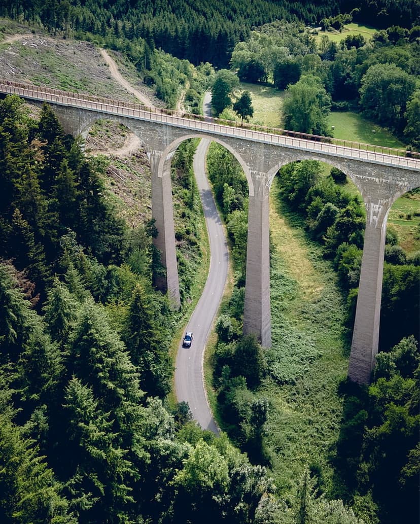 Stone railway viaduct with multiple arches towers over a winding forest road with a white car in a green valley.