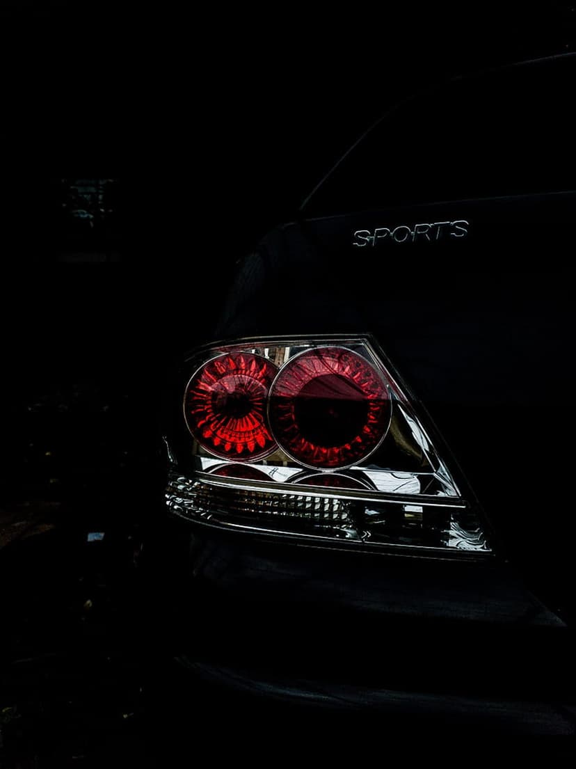 Glowing red taillights on a black car trunk featuring a chrome sports badge in a dark, detailed close-up shot.