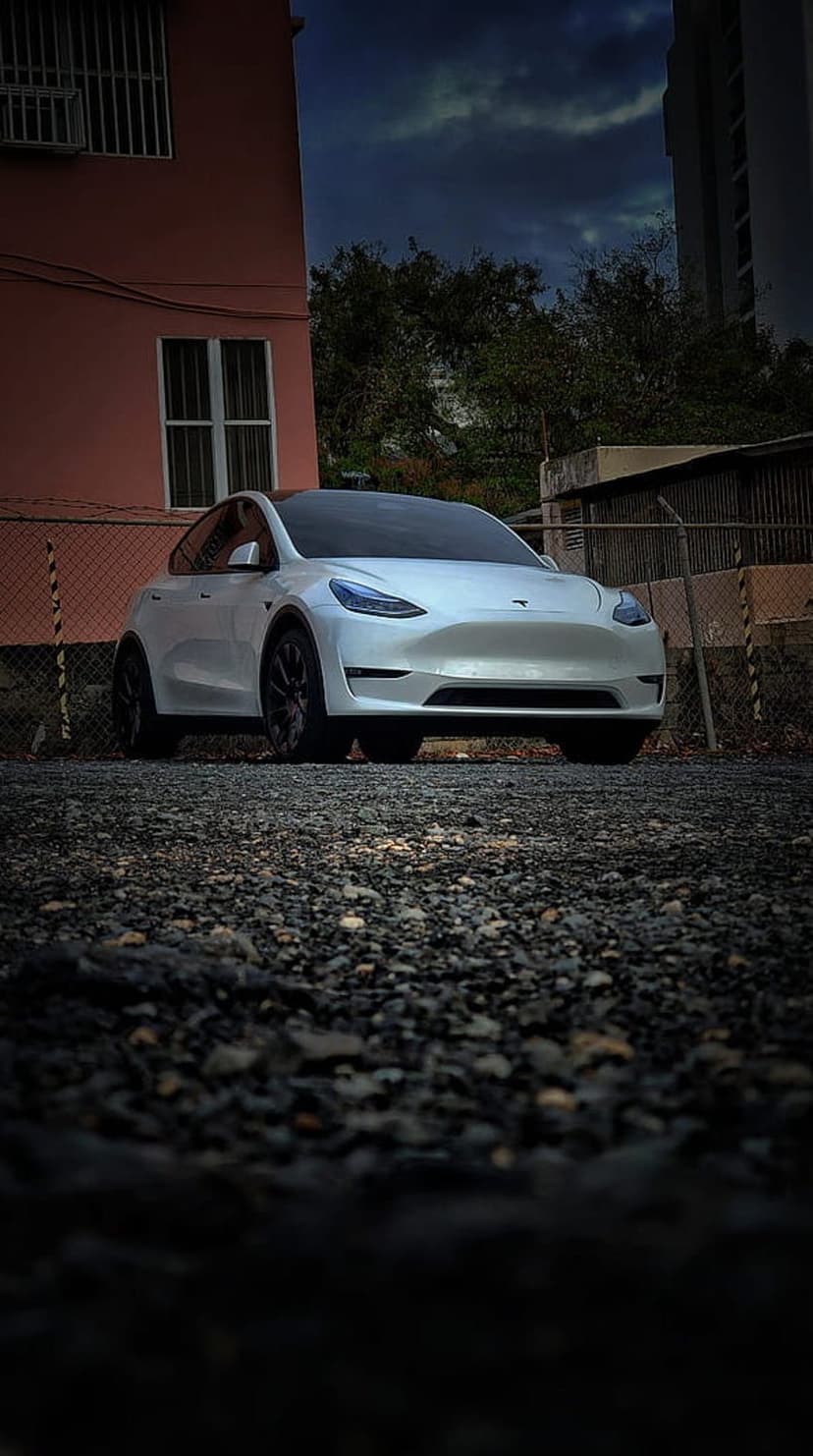White Tesla Model Y parked on a gravel surface before a pink building under a dark, dramatic cloudy sky.