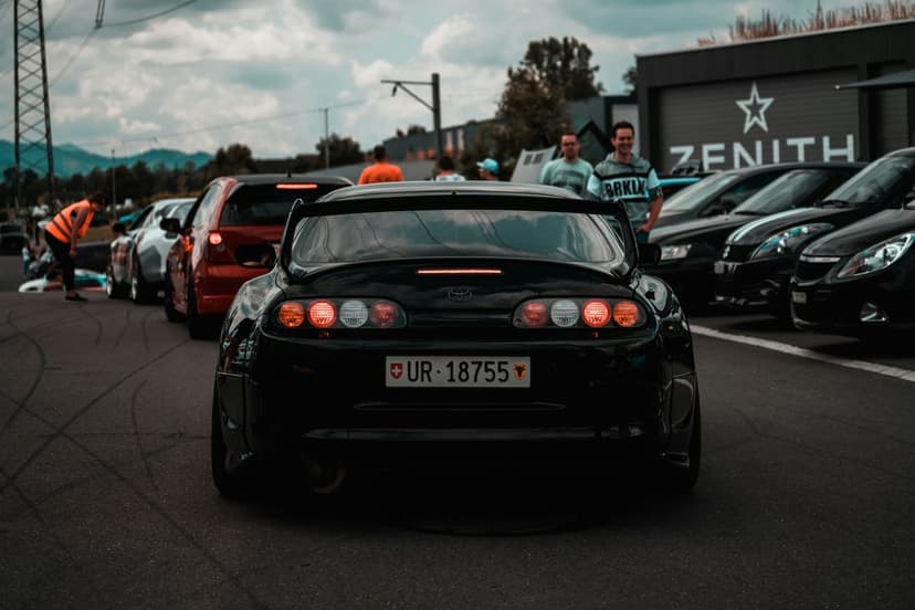 Black Toyota Supra sports car with glowing red LED taillights parked in a row of vehicles at an evening car show.