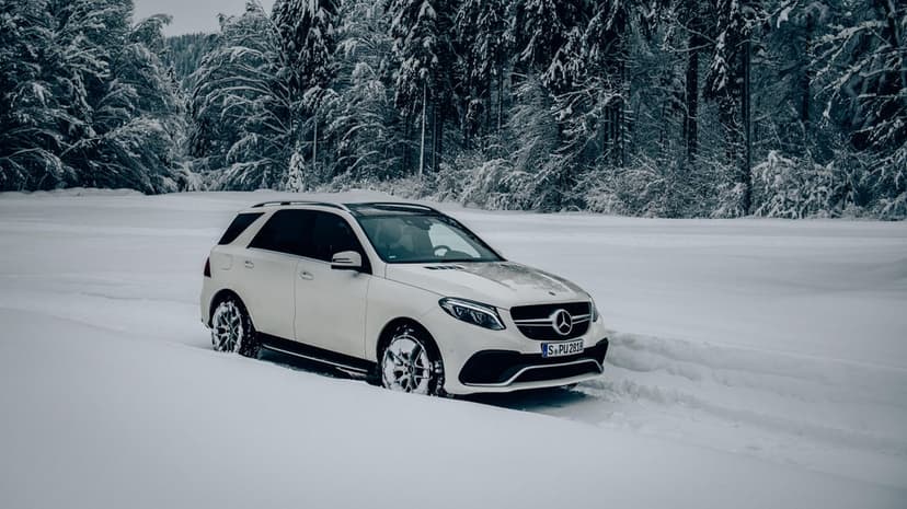 White Mercedes-Benz SUV driving through deep snow tracks in front of a dense frost-covered evergreen forest.