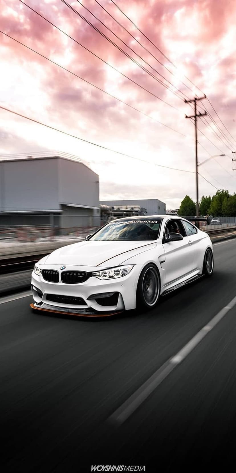 White BMW M4 coupe with custom body kit cruising an urban road beneath a pink and orange cloudy sunset sky.