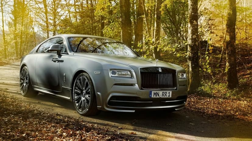 Matte grey Rolls-Royce Wraith on a leaf-strewn road in an autumn forest with warm golden sunlight through trees.