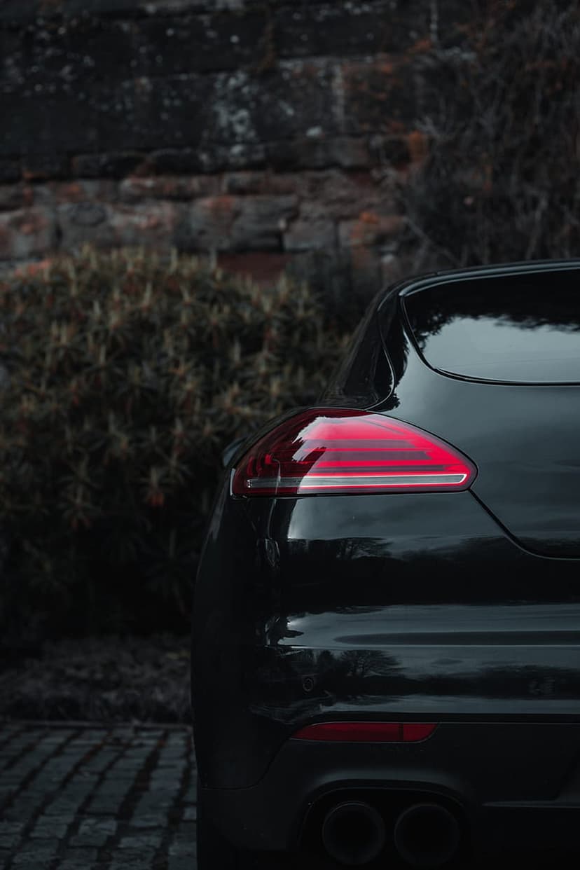 Porsche Panamera rear taillight glowing red on a cobblestone path against a blurred brick wall and foliage.