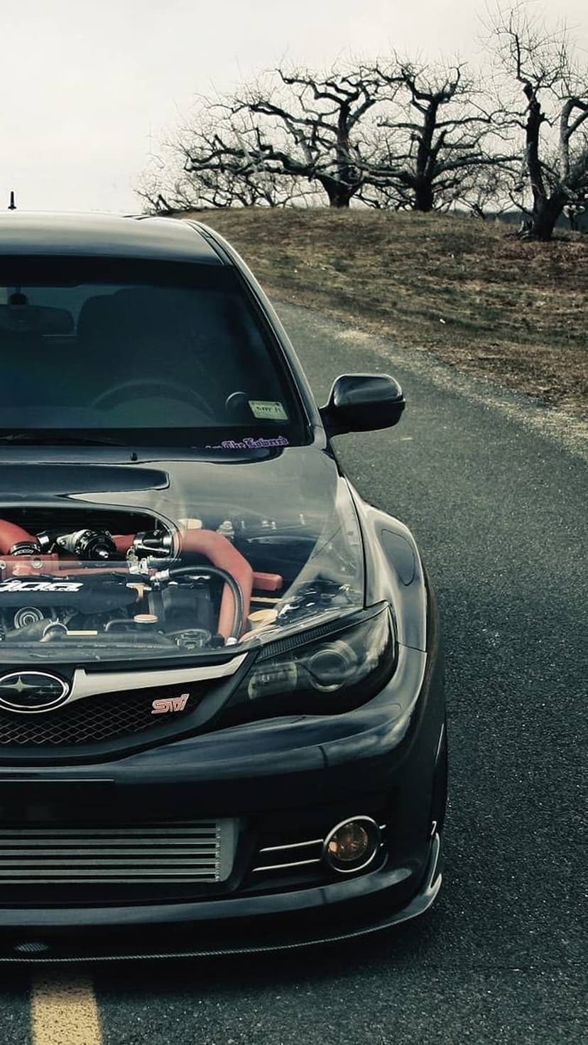 Subaru WRX STI with a transparent hood exposing a modified engine with red piping on a cloudy roadside.