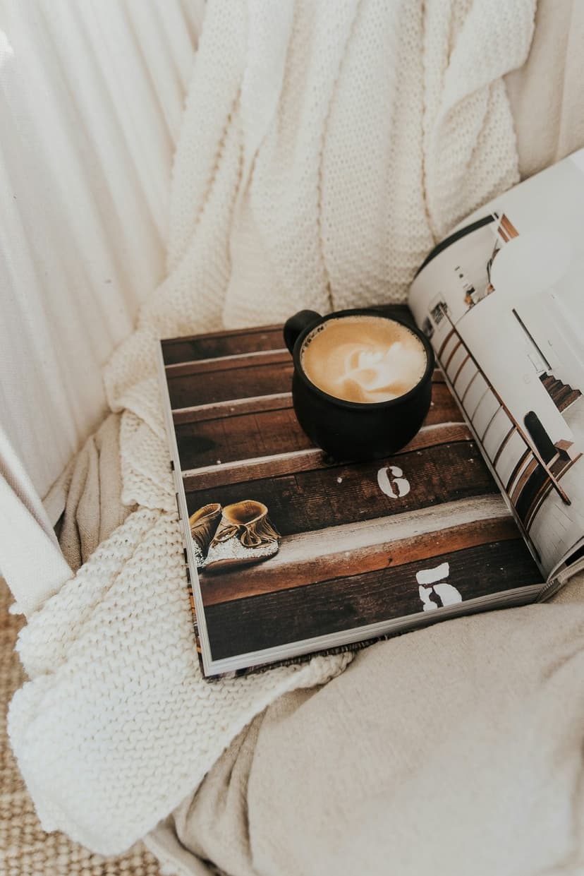 Ceramic coffee cup with latte art on a rustic magazine and textured cream blanket in a cozy vertical scene.