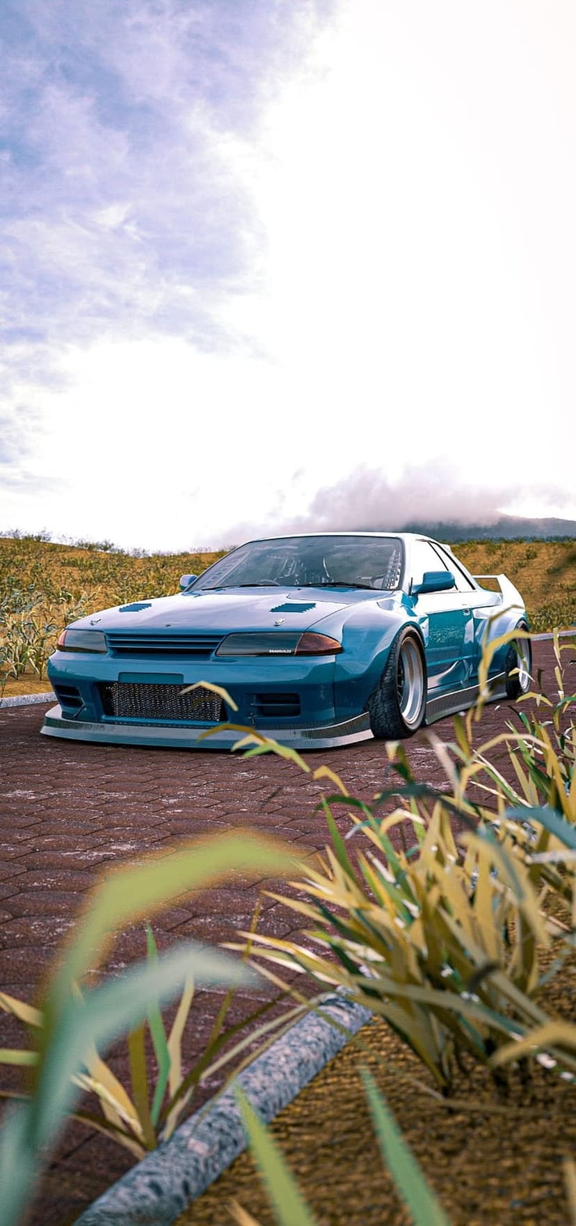 Nissan Skyline R32 with a blue widebody kit parked on bricks before rolling hills and a cloudy mountain sky.