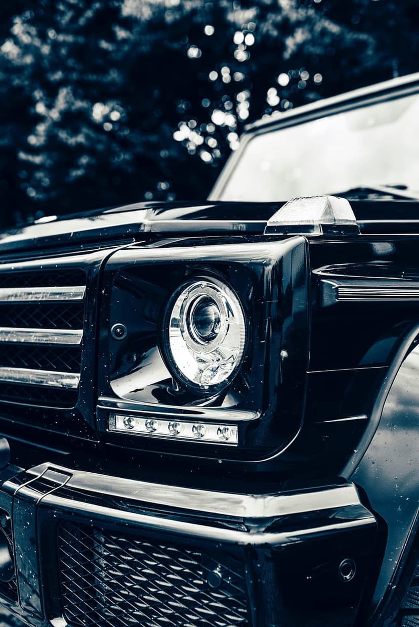 Mercedes G-Class round LED headlight and chrome grille slats against a dark car body and blurred green leaves.