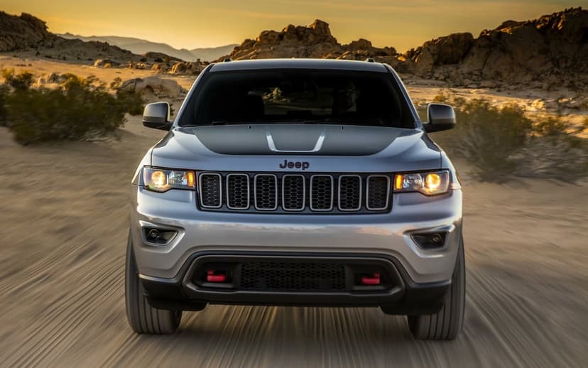 Front view of a silver Jeep Grand Cherokee driving through a rocky desert under a warm golden sunset sky.