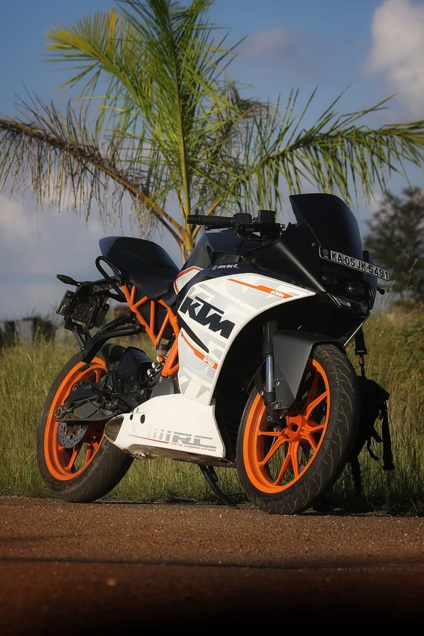KTM RC motorcycle with orange wheels parked on green grass under a palm tree against a clear blue sky.