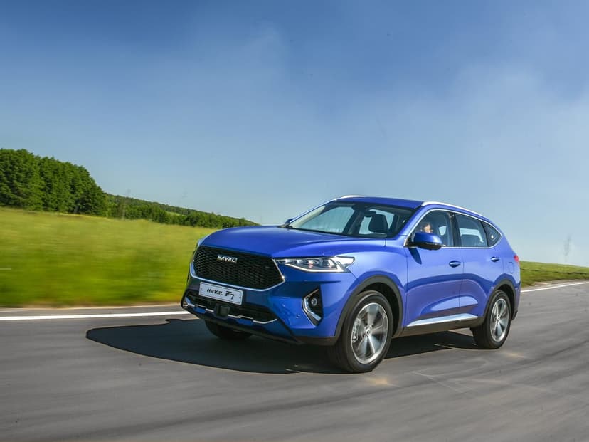 Blue Haval F7 SUV driving fast on a paved road past a green field and trees under a vast clear blue sky.