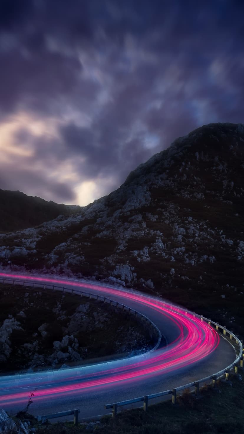 Neon pink and blue light trails wind around a dark mountain road beneath a cloudy night sky on a vertical screen.