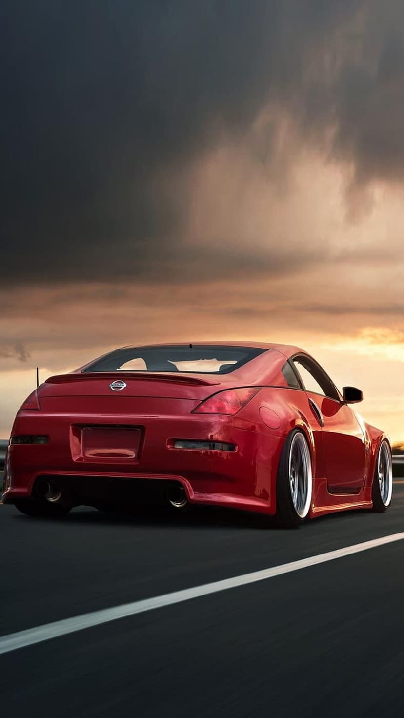 Red Nissan 350Z sports car driving on a highway during a cloudy sunset with golden light and custom wheels.