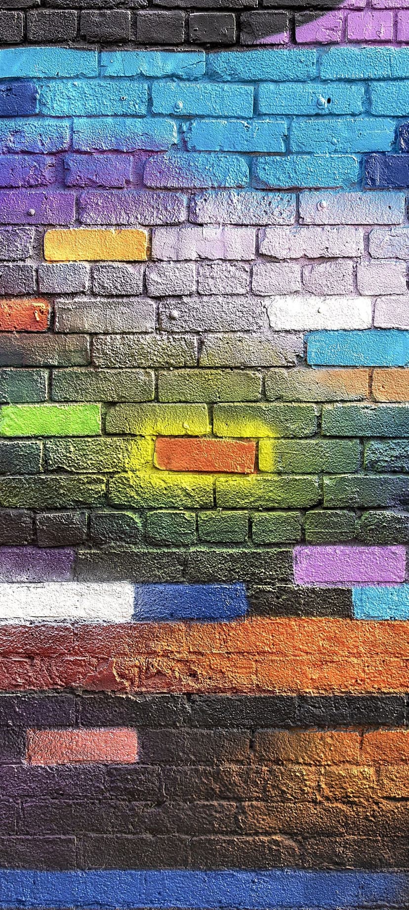 Multicolored horizontal graffiti stripes with spray paint splatters cover a textured brick wall surface.