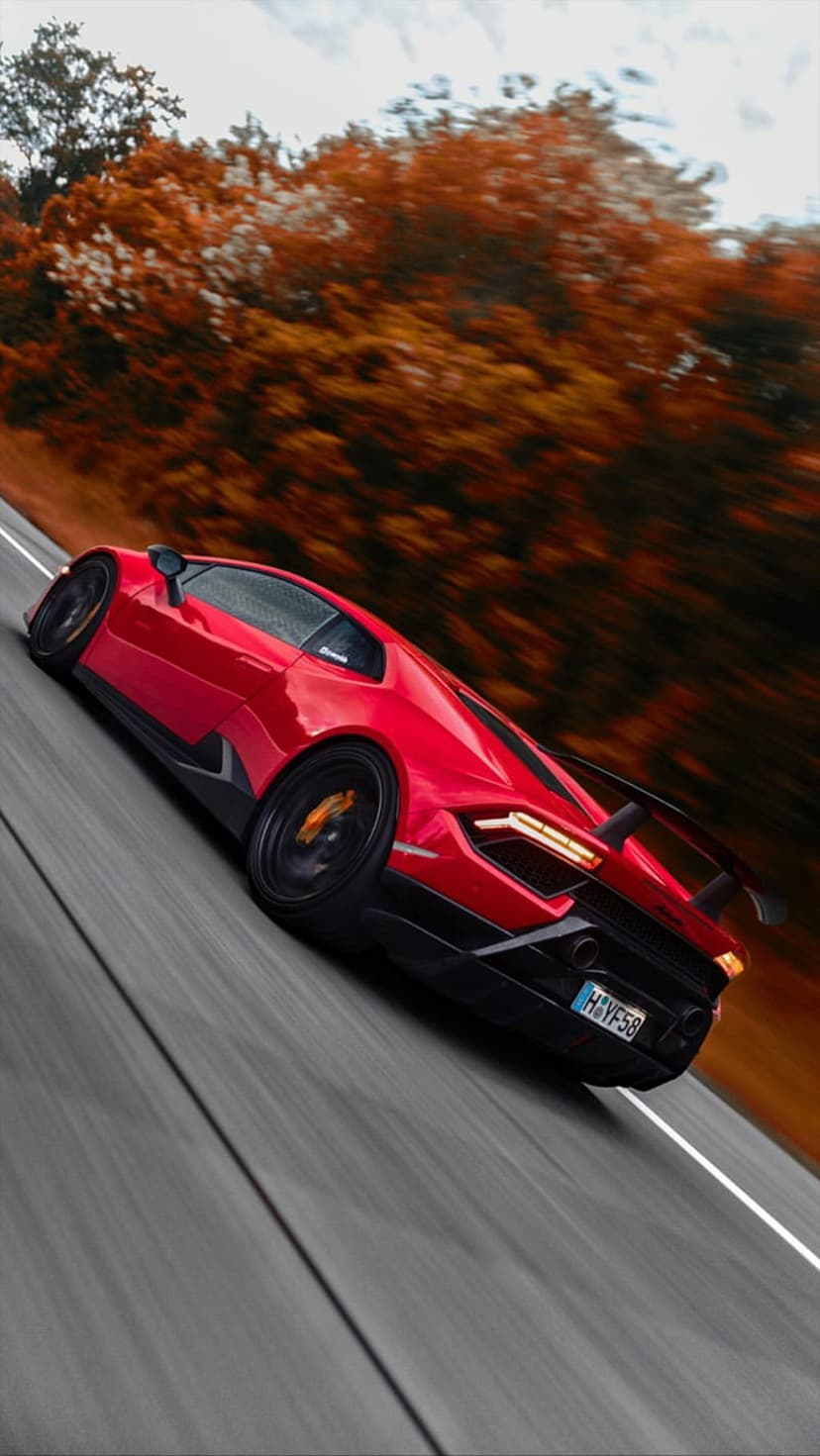 Red sports car racing on a blurred autumn road with glowing taillights and golden forest foliage in the distance.