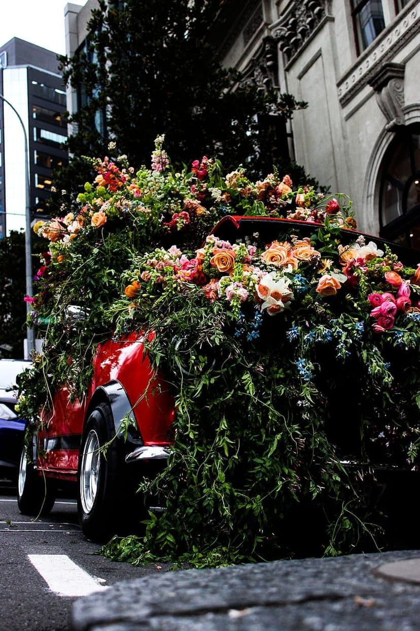 Red car covered in thick layers of blooming flowers and green vines on a city street with urban buildings.