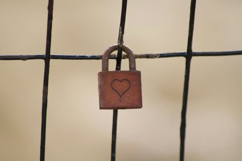 Rusty padlock with a small etched heart hangs on a dark chain-link fence against a soft-focus background.