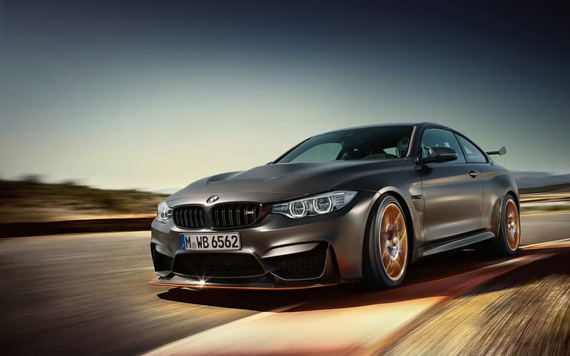 Dark grey BMW M4 GTS with orange accents racing on a track with motion blur under a clear sky.