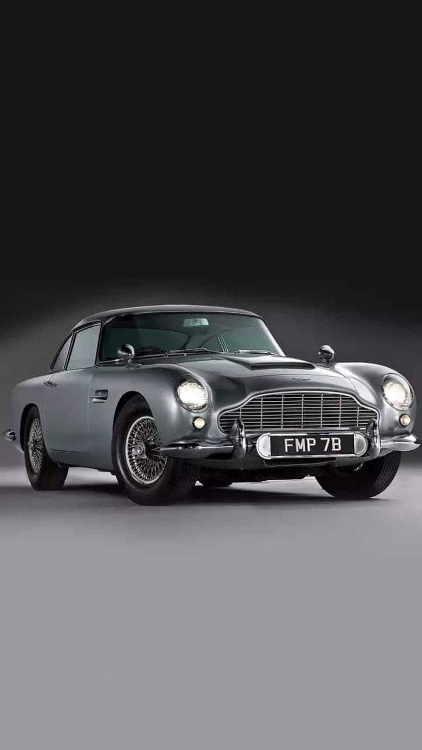 Silver Aston Martin DB5 classic car with wire wheels and chrome grille parked in a dark studio setting.