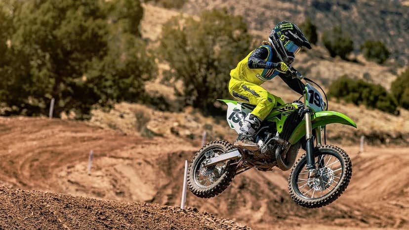 Motocross rider in yellow gear jumps a green Kawasaki dirt bike over a brown track against a hilly backdrop.