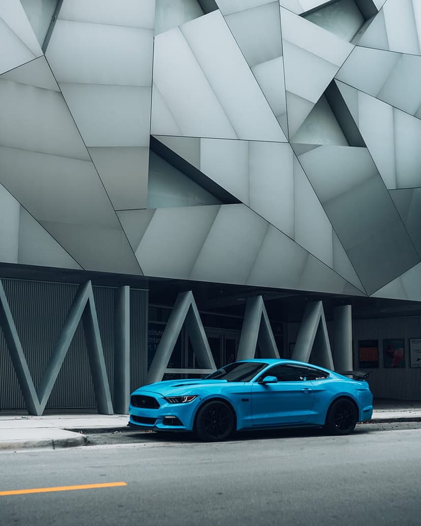 Blue Ford Mustang parked on an urban street in front of a modern building with gray geometric panels.