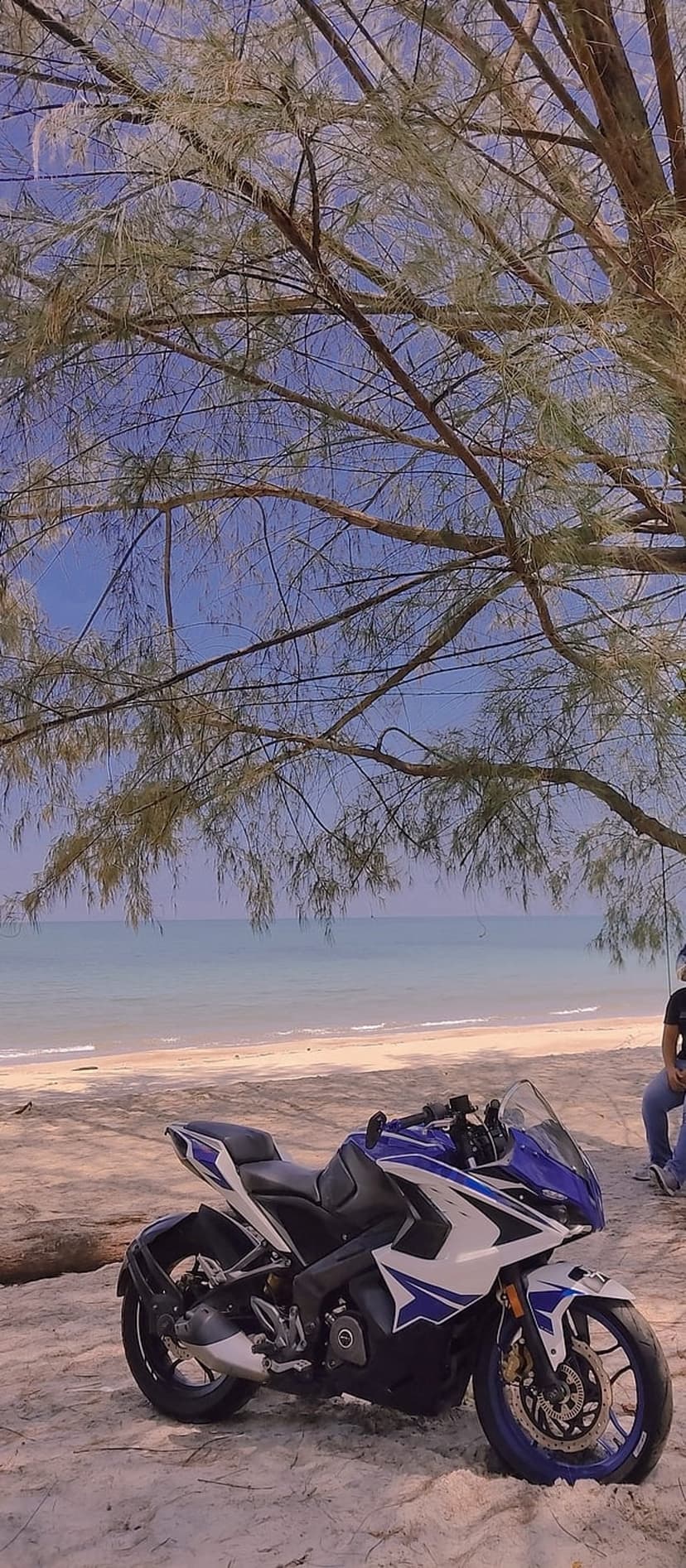 Blue and white sports motorcycle parked on a sandy beach with the ocean and a clear sky in the background.