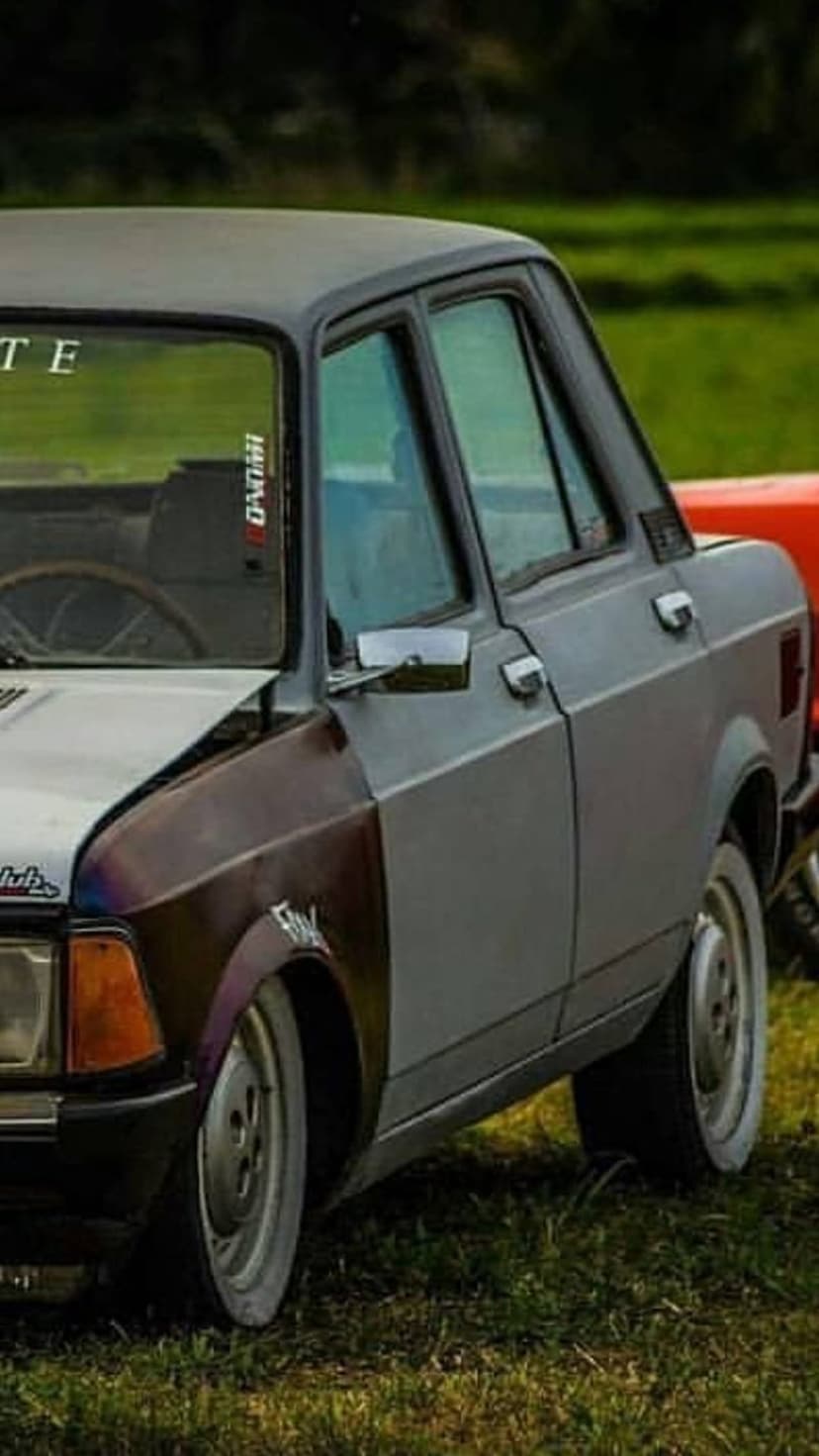 Vintage sedan with a rusty brown fender and grey body parked on green grass shows off white-wall tires.