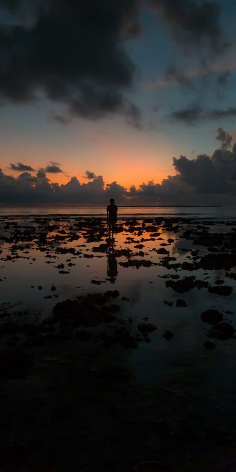 A lone person stands in a tidal pool reflecting a vibrant orange and blue twilight sky over rugged wet rocks.