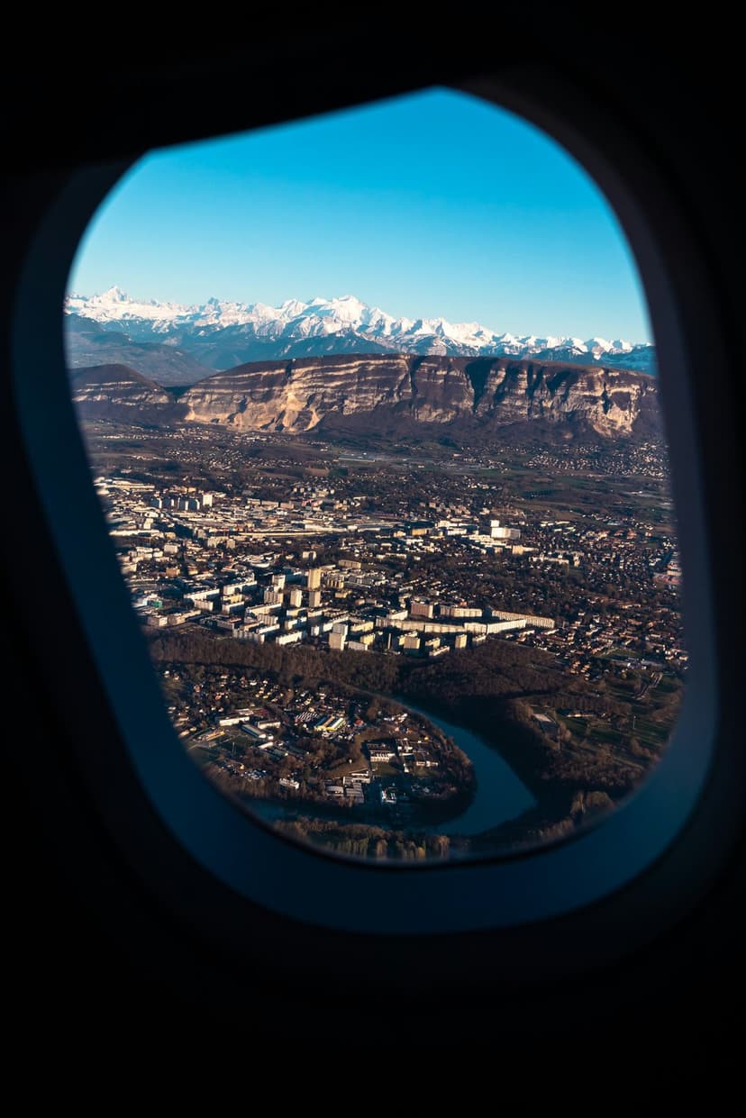 Grenoble city buildings sprawl beside a winding river with massive snow-capped mountains under a clear blue sky.