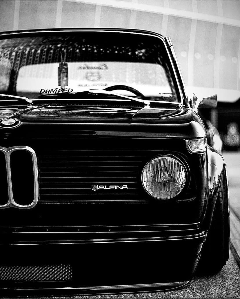 Vintage BMW Alpina front headlight and grille with wet surface reflections in a black and white close-up shot.