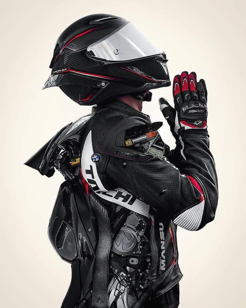 Motorcyclist in red and black racing gear with hands clasped over a glossy helmet against a neutral background.
