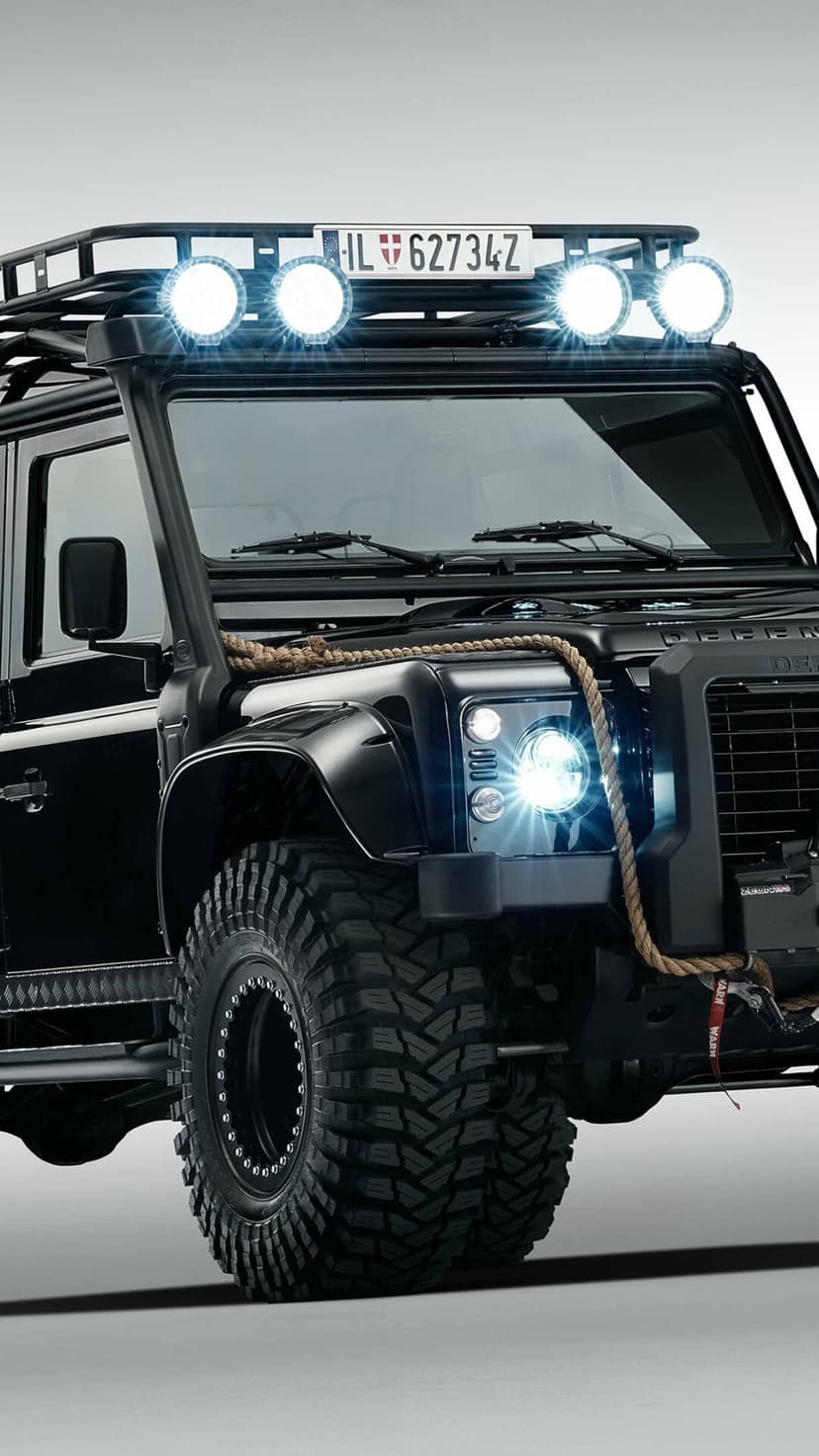 Black Land Rover Defender with glowing roof lights and massive off-road tires on a light background.