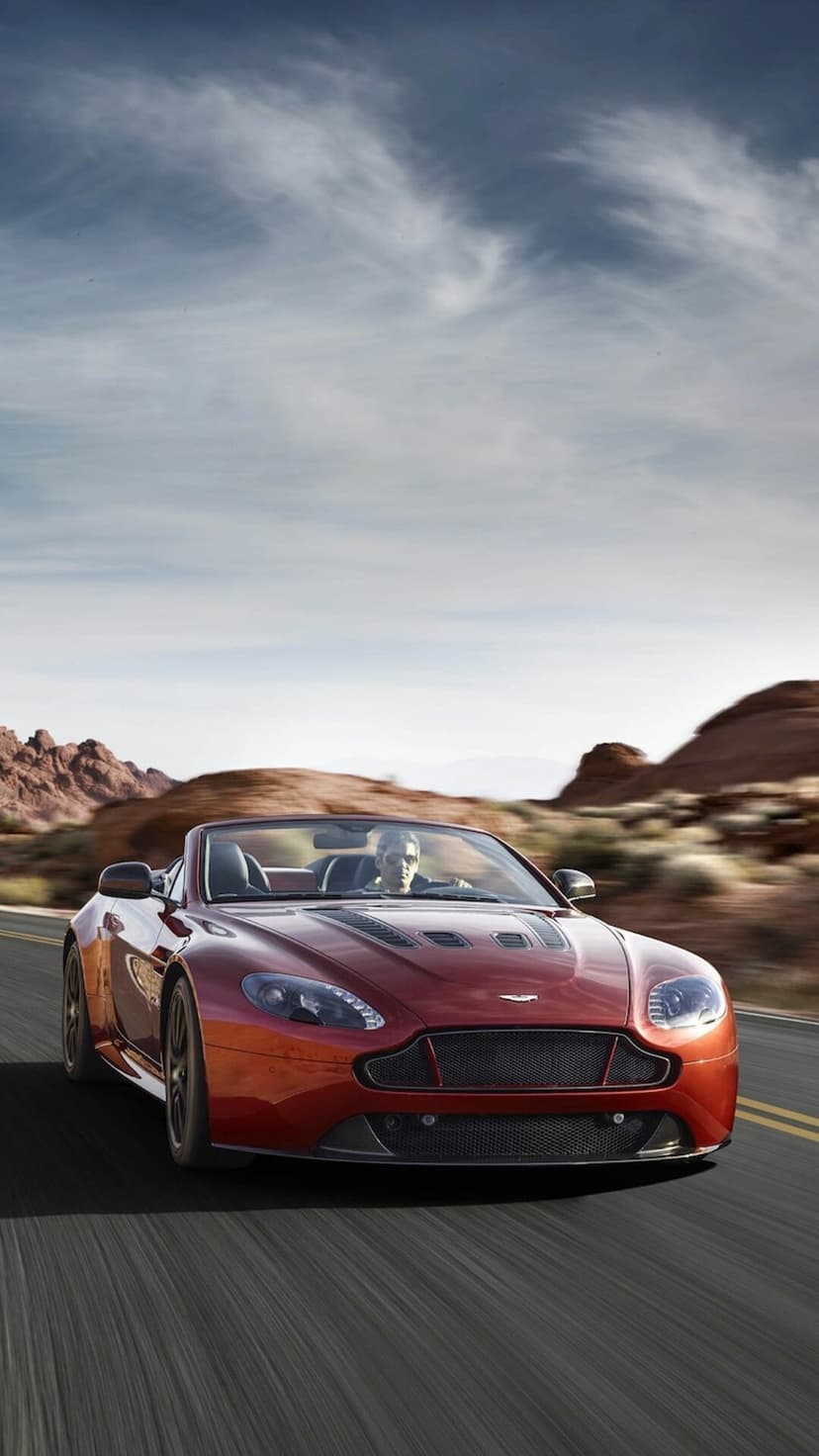 Red Aston Martin convertible speeding through a desert canyon with red rock formations under a blue sky.