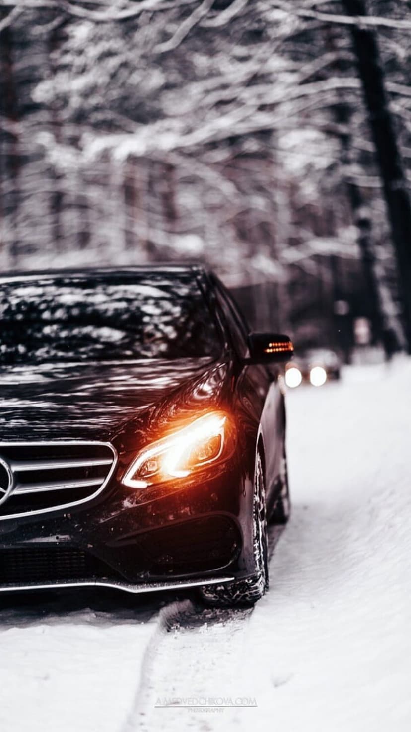 Black Mercedes sedan with glowing headlights drives on a narrow road between snow-covered evergreen trees.